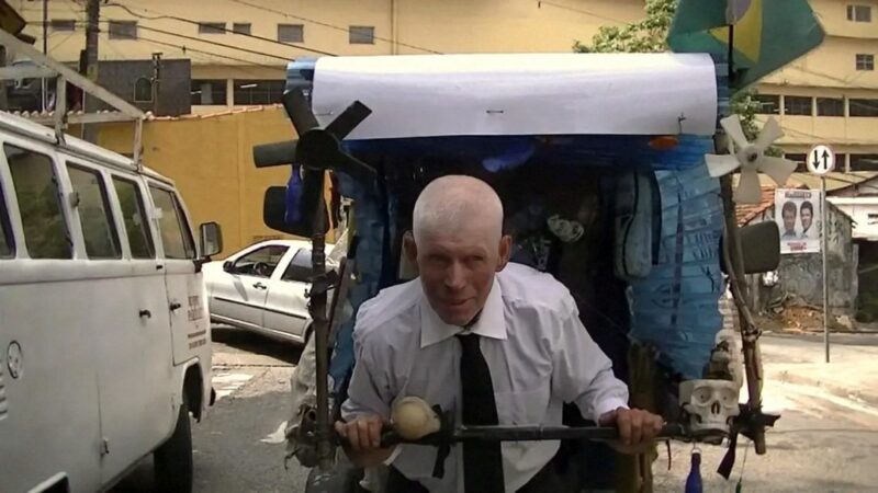 iwan&han A man in a white shirt and black tie pushes a blue vehicle decorated with small flags on a street with cars and buildings in the background. He appears focused, with an intense expression.