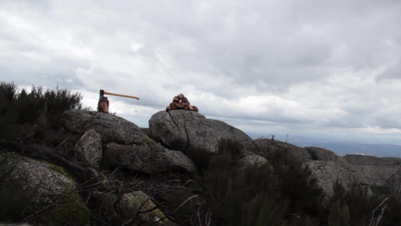 iwan&han A cloudy sky over a rocky landscape with large boulders. An axe is embedded in one of the rocks, and a small pile of rocks is stacked nearby. Sparse vegetation surrounds the area.