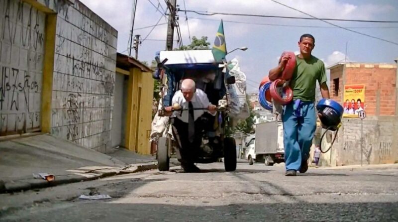 iwan&han A man pushes a cart filled with various items down a narrow, graffiti-covered street. Another man walks beside him, carrying coiled ropes in each hand. A Brazilian flag is visible on the cart. The sky is partly cloudy.