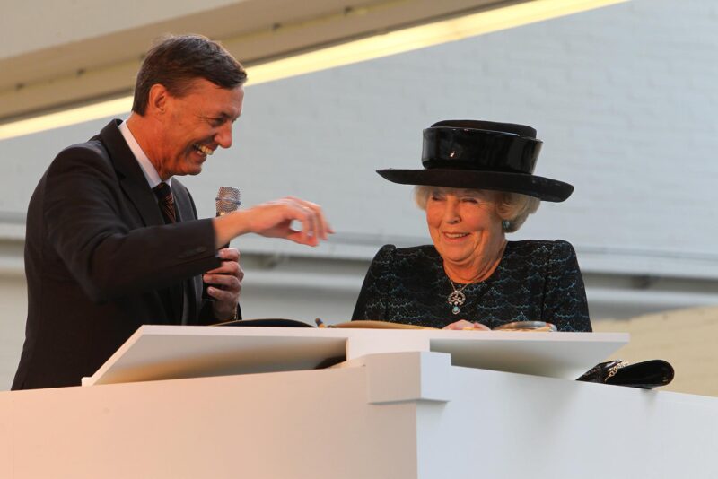 iwan&han A smiling man in a suit holds a microphone while gesturing toward an older woman wearing a large black hat and dark dress, who is signing a book at a white podium indoors.