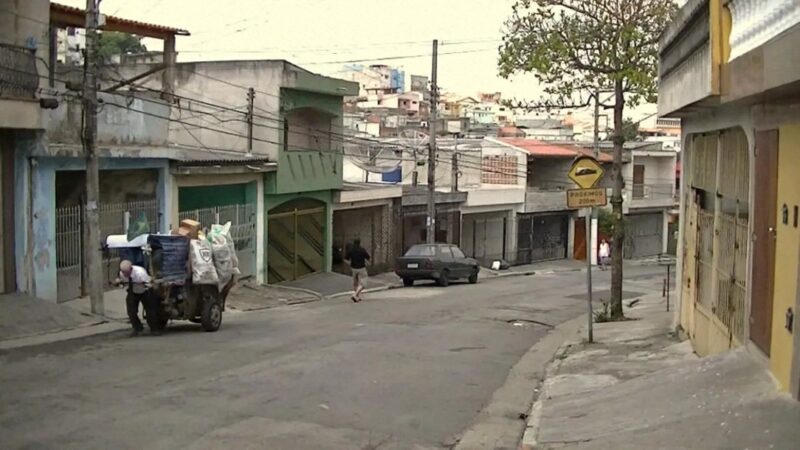 iwan&han A quiet, slightly uphill street in an urban neighborhood features a person pushing a cart loaded with bags. Buildings line both sides, and a small car is parked on the right. A tree stands next to a street sign. There’s a hilly area in the background.
