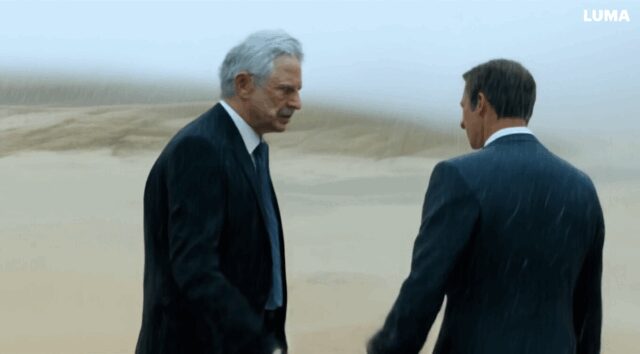 man with gray hair wearing a suit, walks in the dessert on a rainy day and meet his doppelgänger with grey hair and then they shake hands-fcd410