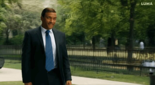 Man with a suit walks in the park, during his short walk he passes another man, they both stop and then they look at each other for 3 seconds. Than they shake hands and walk away-98b363